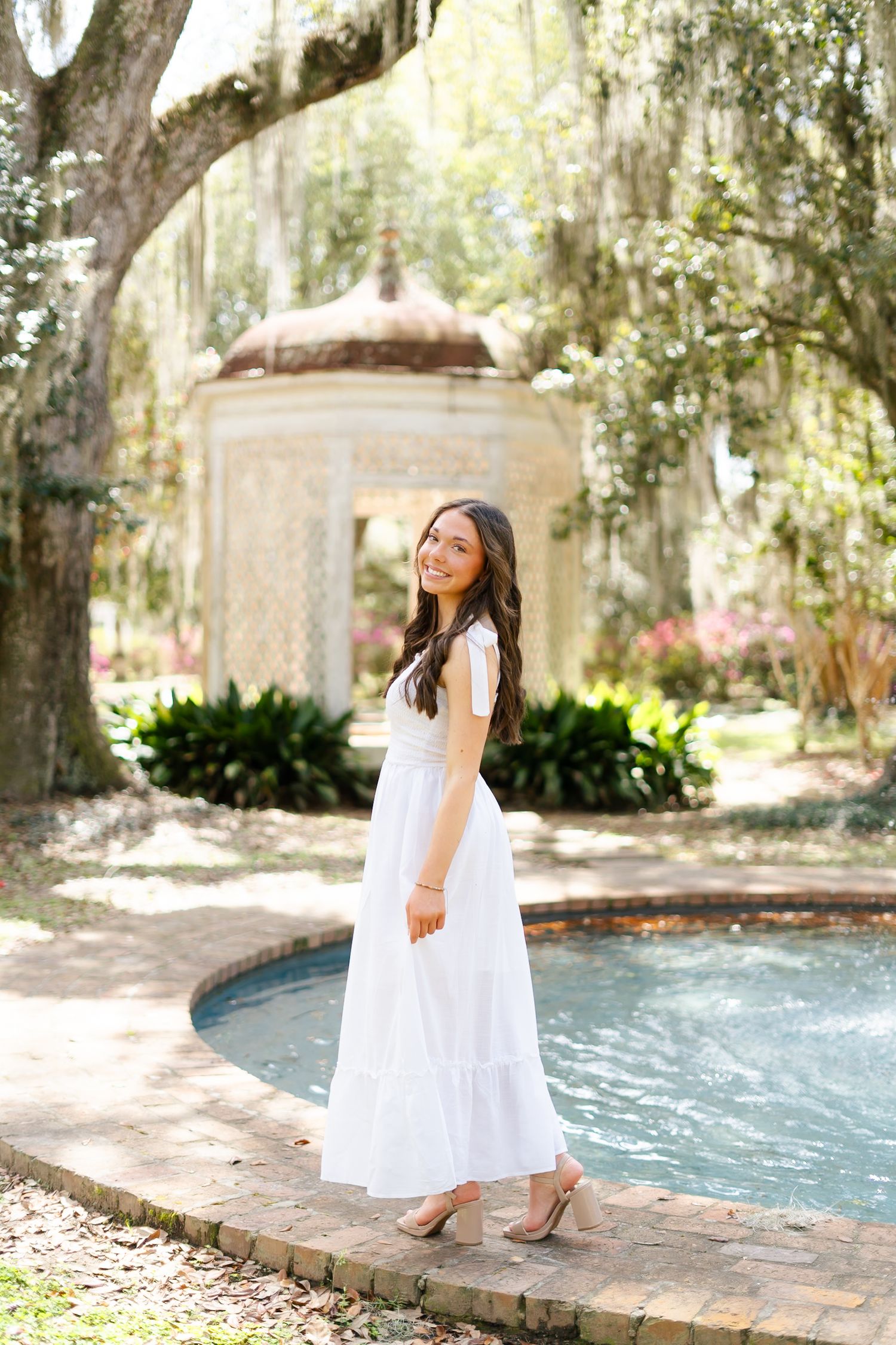 Woman in flowing white dress stands by ornate garden fountain under Spanish moss draped trees in romantic setting.