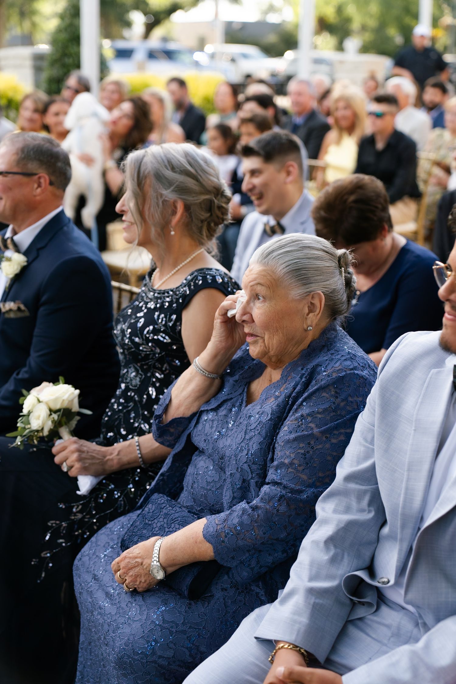 Wedding guests seated in formal attire watch ceremony unfold outdoors.