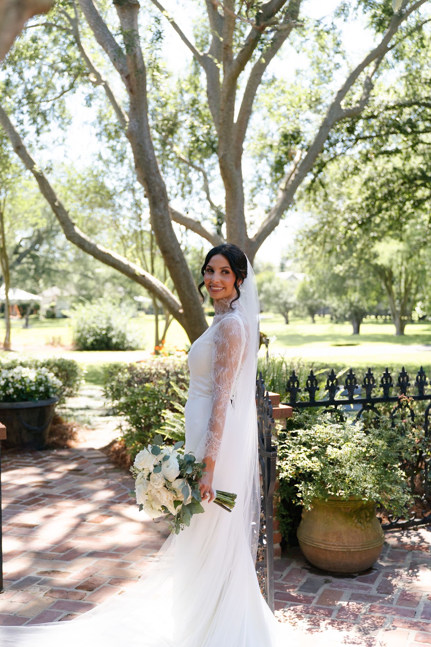 Bride in elegant white dress poses in garden setting with lush trees and brick pathways.