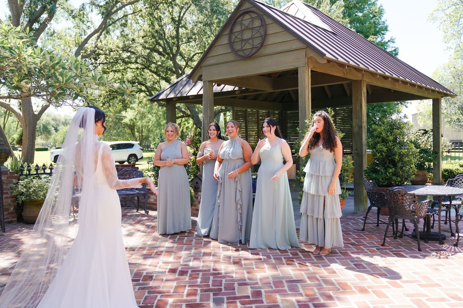 Bridal party in soft blue gowns gathers on a brick patio under a wooden pavilion during an outdoor celebration.