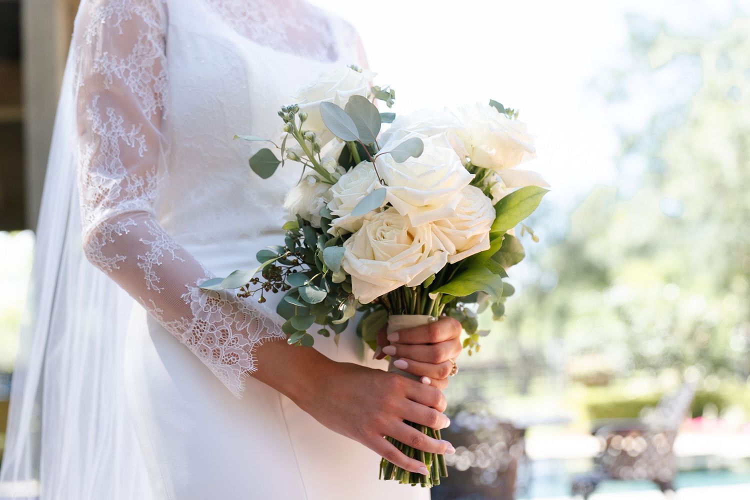 Bride holding a beautiful white and peach rose bouquet with lace sleeve details in soft natural lighting.
