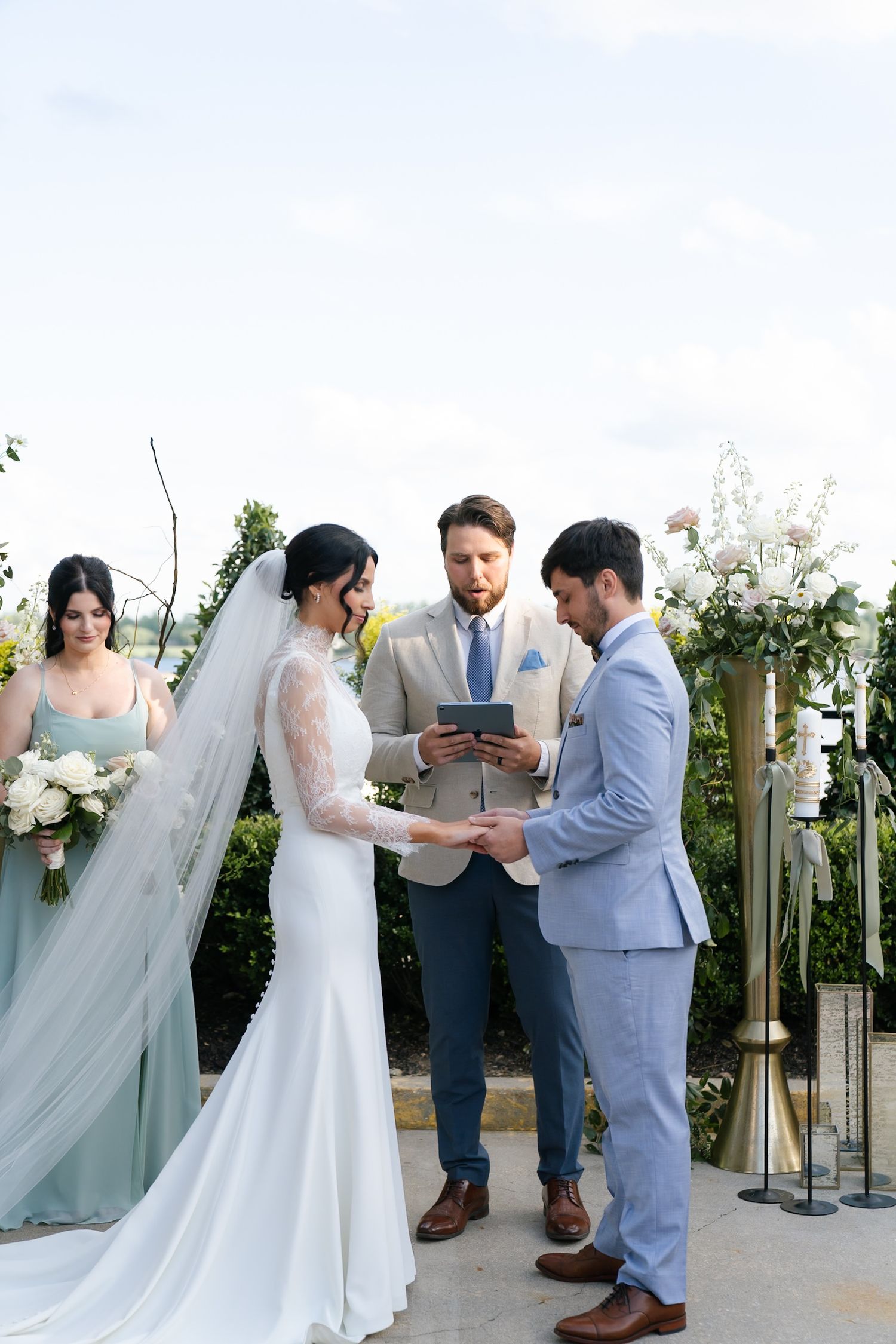 Wedding party gathers for outdoor ceremony with white floral arrangements and flowing sage green dresses.