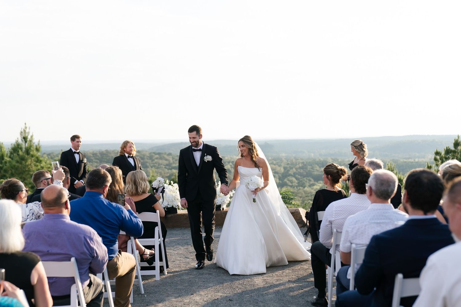A wedding ceremony takes place on a scenic overlook with guests seated and mountains visible in the distance.