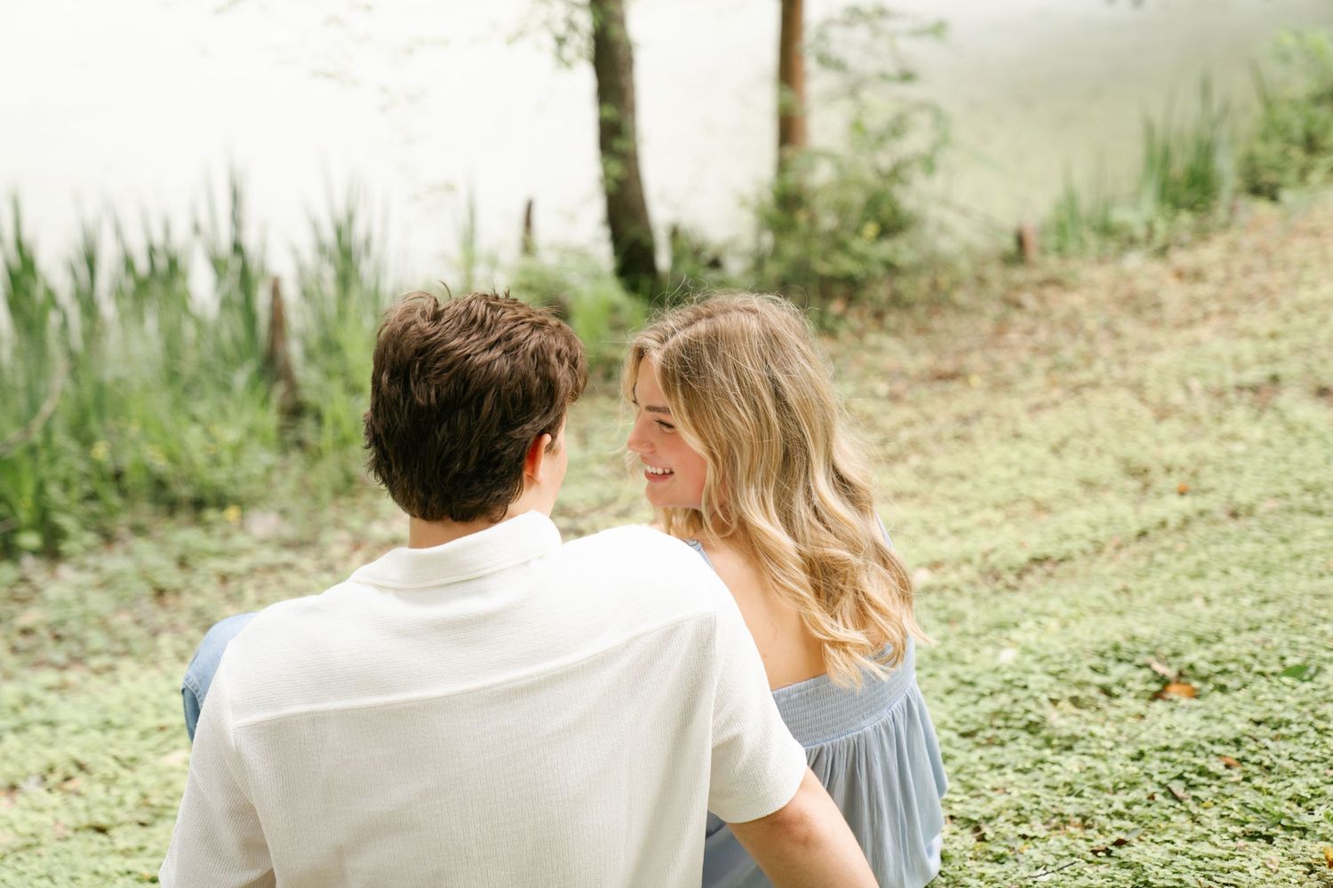 A couple sits together outdoors on a grassy hillside during what appears to be an engagement photo session.