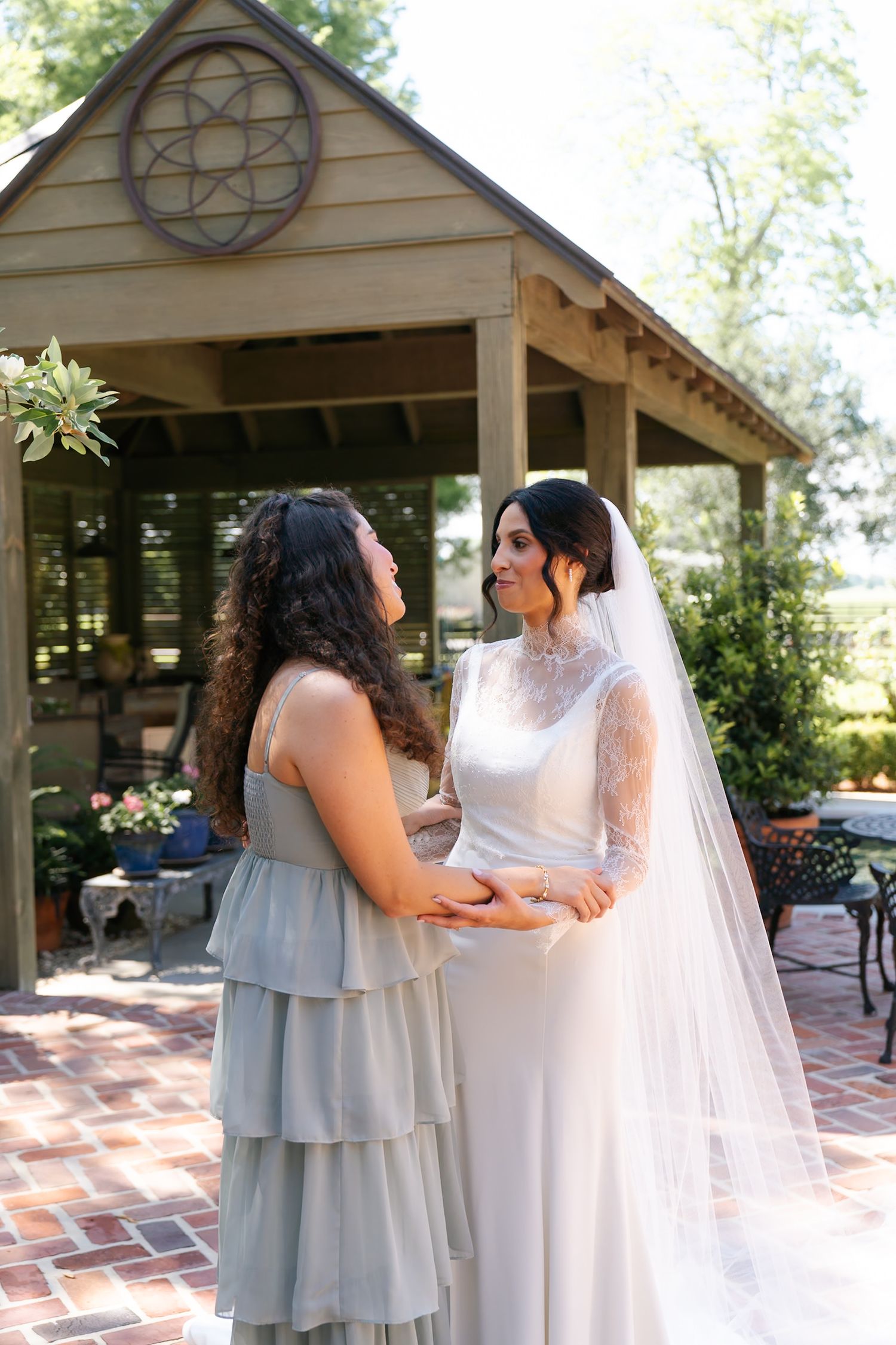Wedding attendants in gray dresses help adjust a bride's flowing white gown and veil on a brick patio.