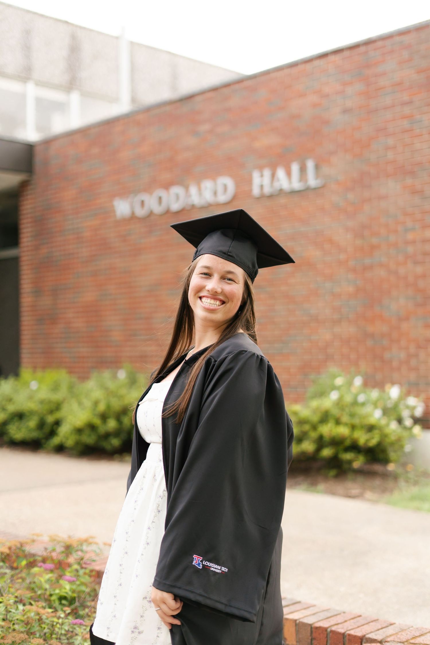 Graduate in black cap and gown poses for portraits in front of brick building during commencement ceremony.