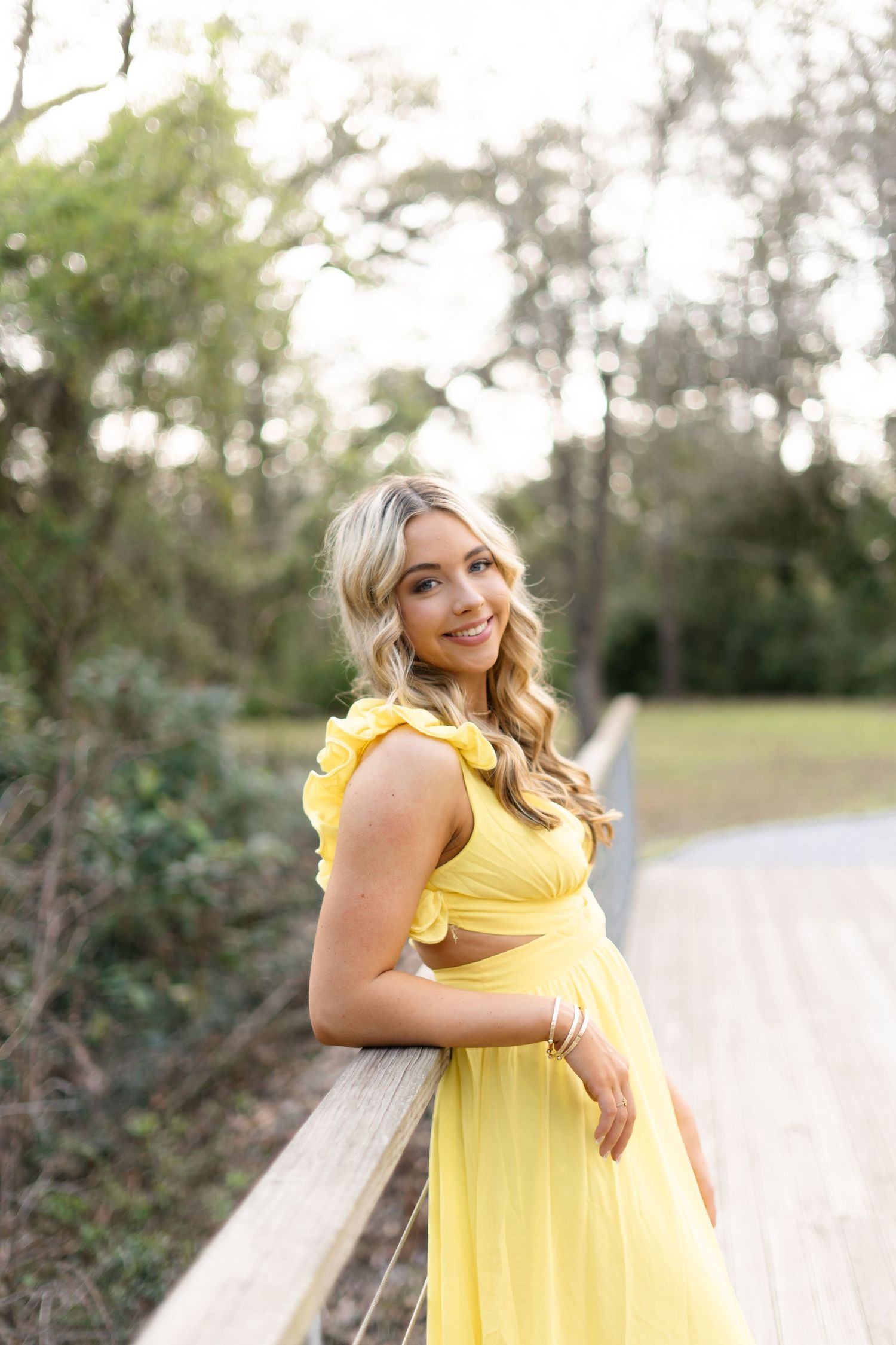 A person in a bright yellow sundress poses outdoors on a wooden bench during a sunny day.