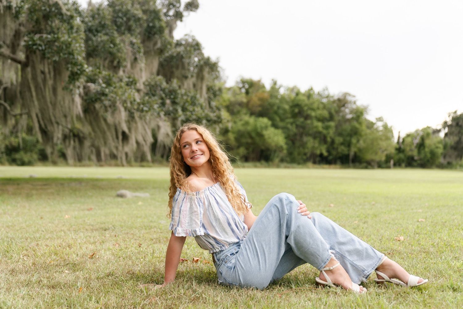 A person in a white off-shoulder top and jeans relaxes on grass under Spanish moss-draped trees in a park.