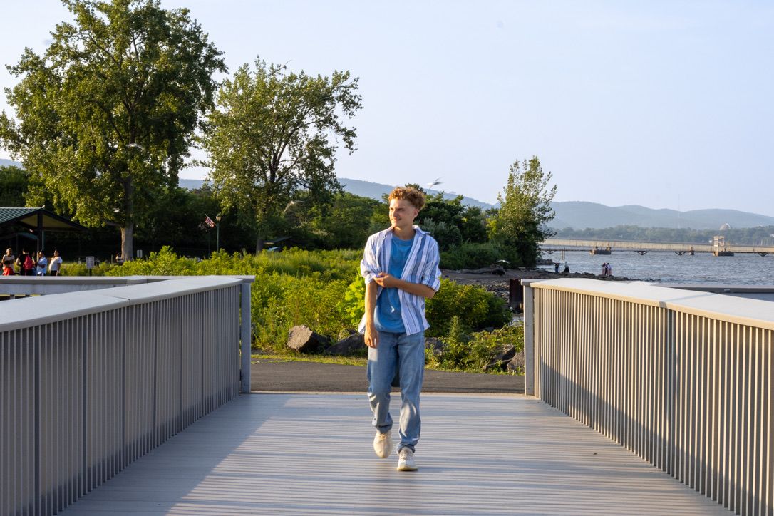 Person in blue shirt walking along scenic waterfront boardwalk with trees and mountains in background.