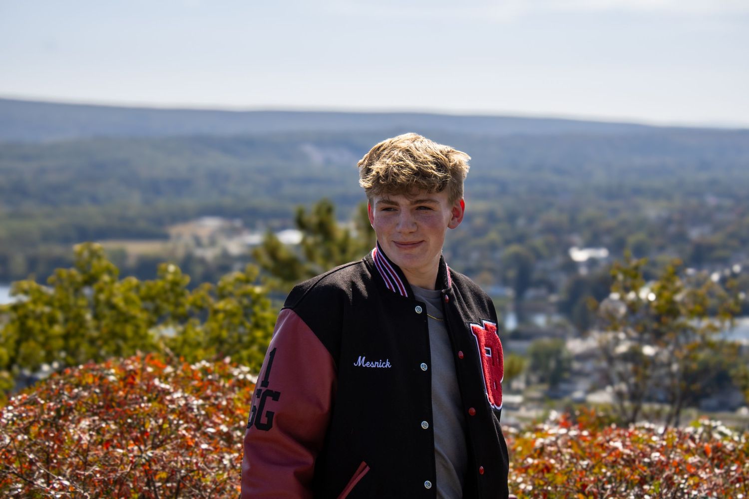 Student in varsity jacket poses with scenic mountain valley view in background.