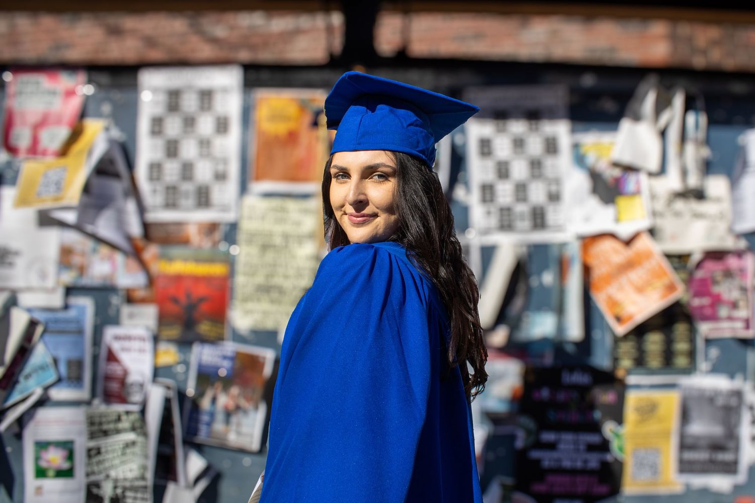 Graduate in blue cap and gown poses against wall of posters.
