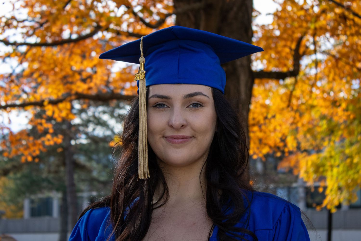 Graduate in blue cap and gown poses against vibrant fall foliage.