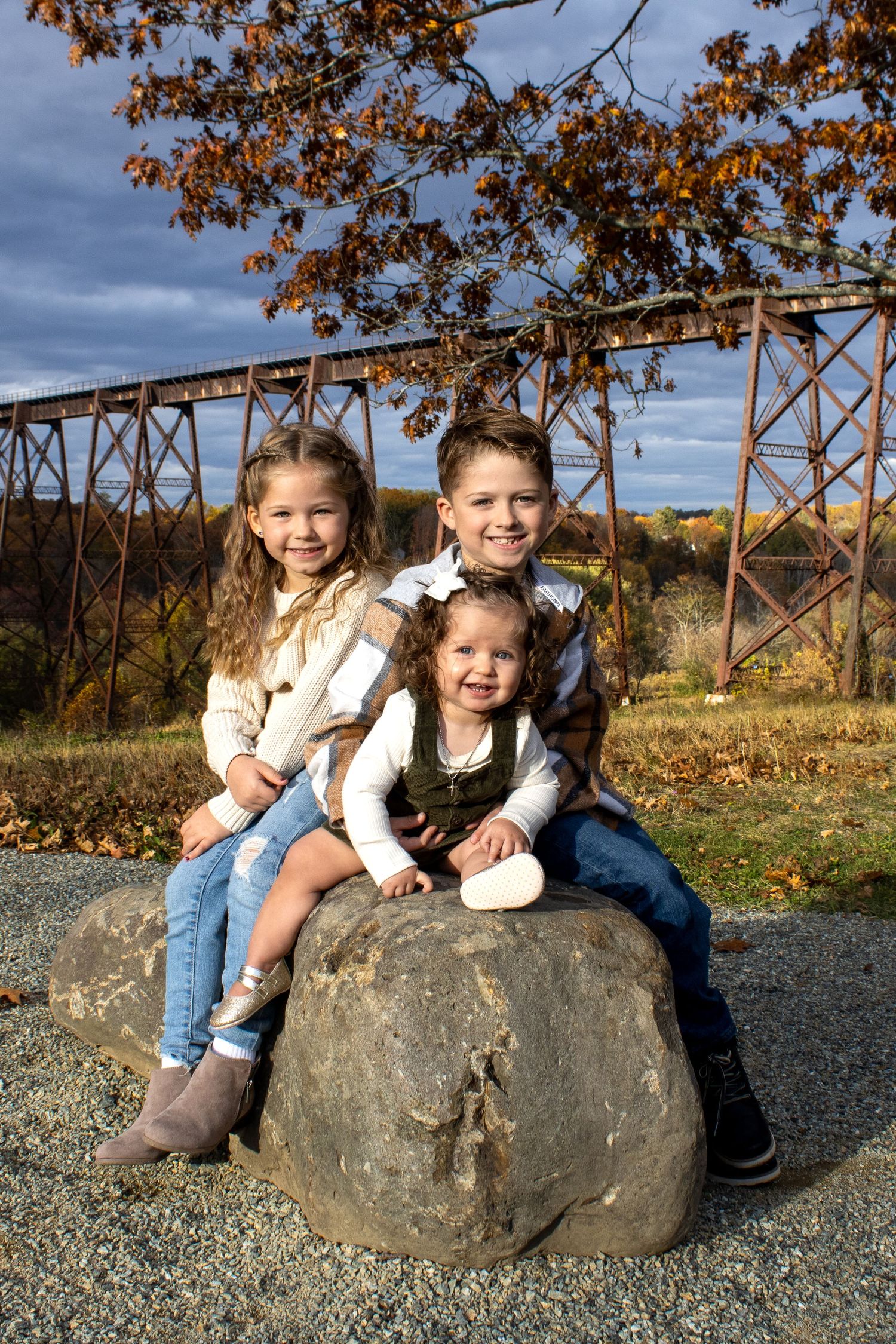 Three children sitting on large boulder with rustic train trestle and fall foliage in background.