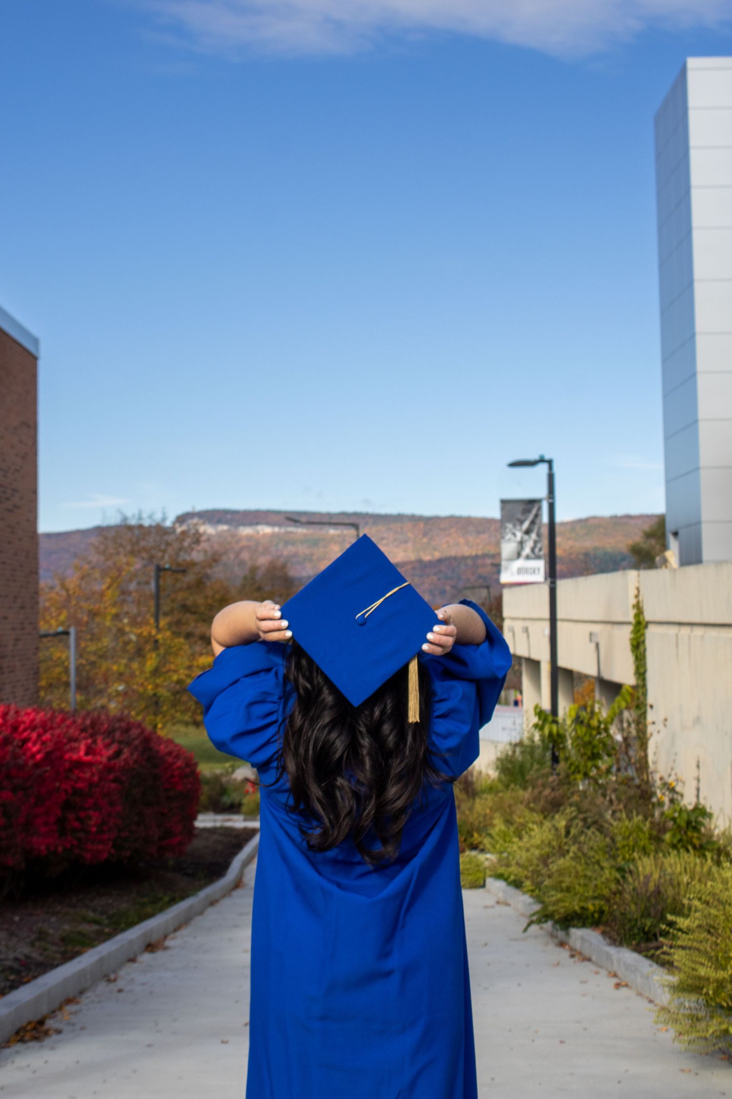 Graduate in blue cap and gown holds up diploma against autumn campus backdrop.