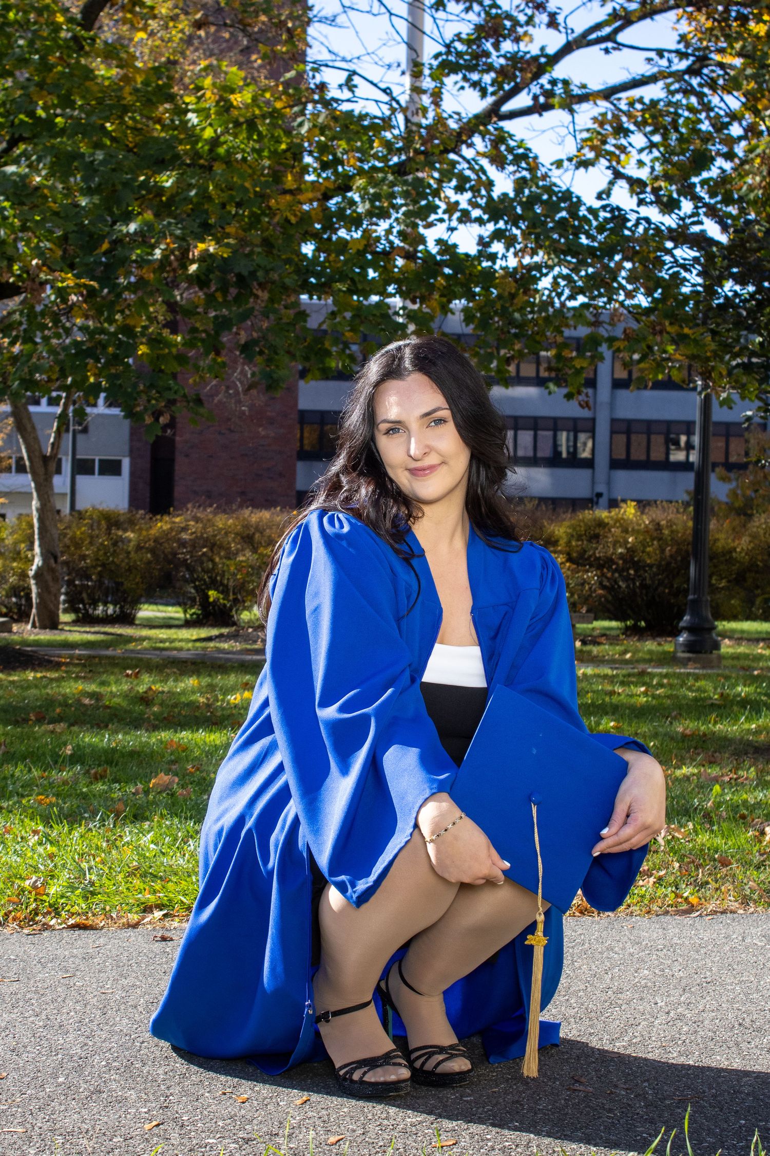 Graduate in blue commencement regalia poses on campus lawn in autumn.