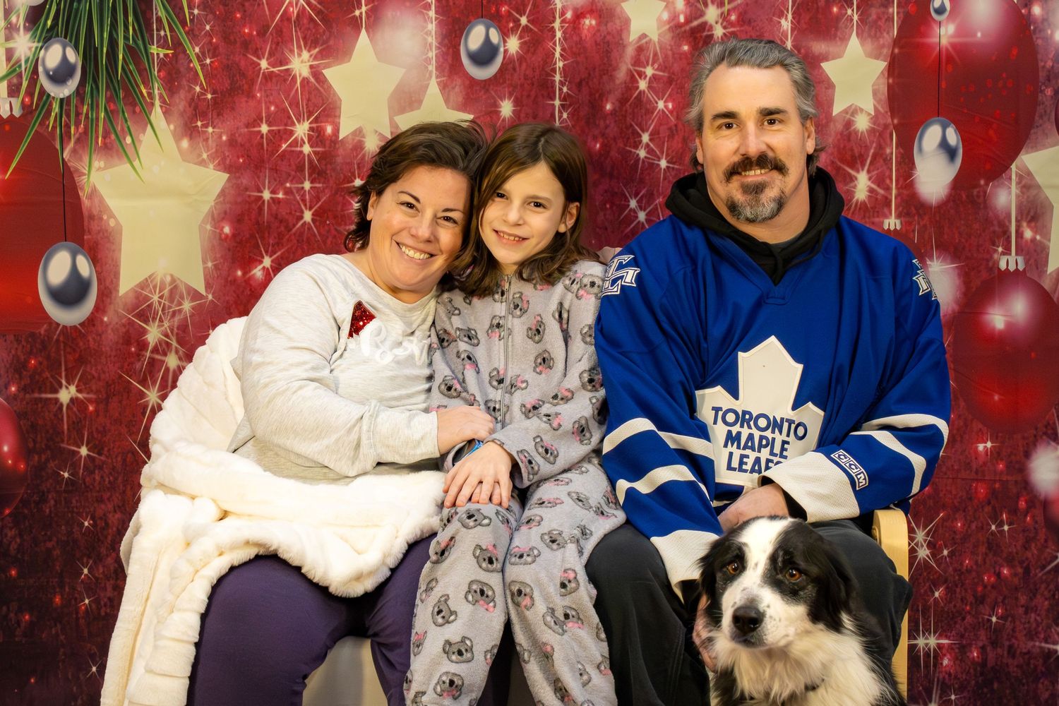 A festive holiday portrait with two adults, a child and a black and white dog against a red sparkly backdrop with ornaments.