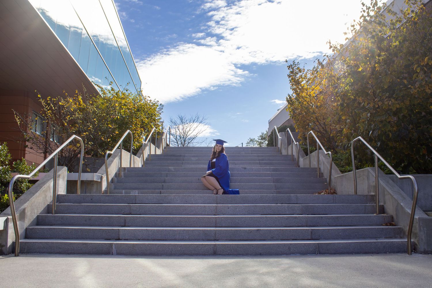 Graduate in blue regalia ascends concrete campus stairs on sunny day.