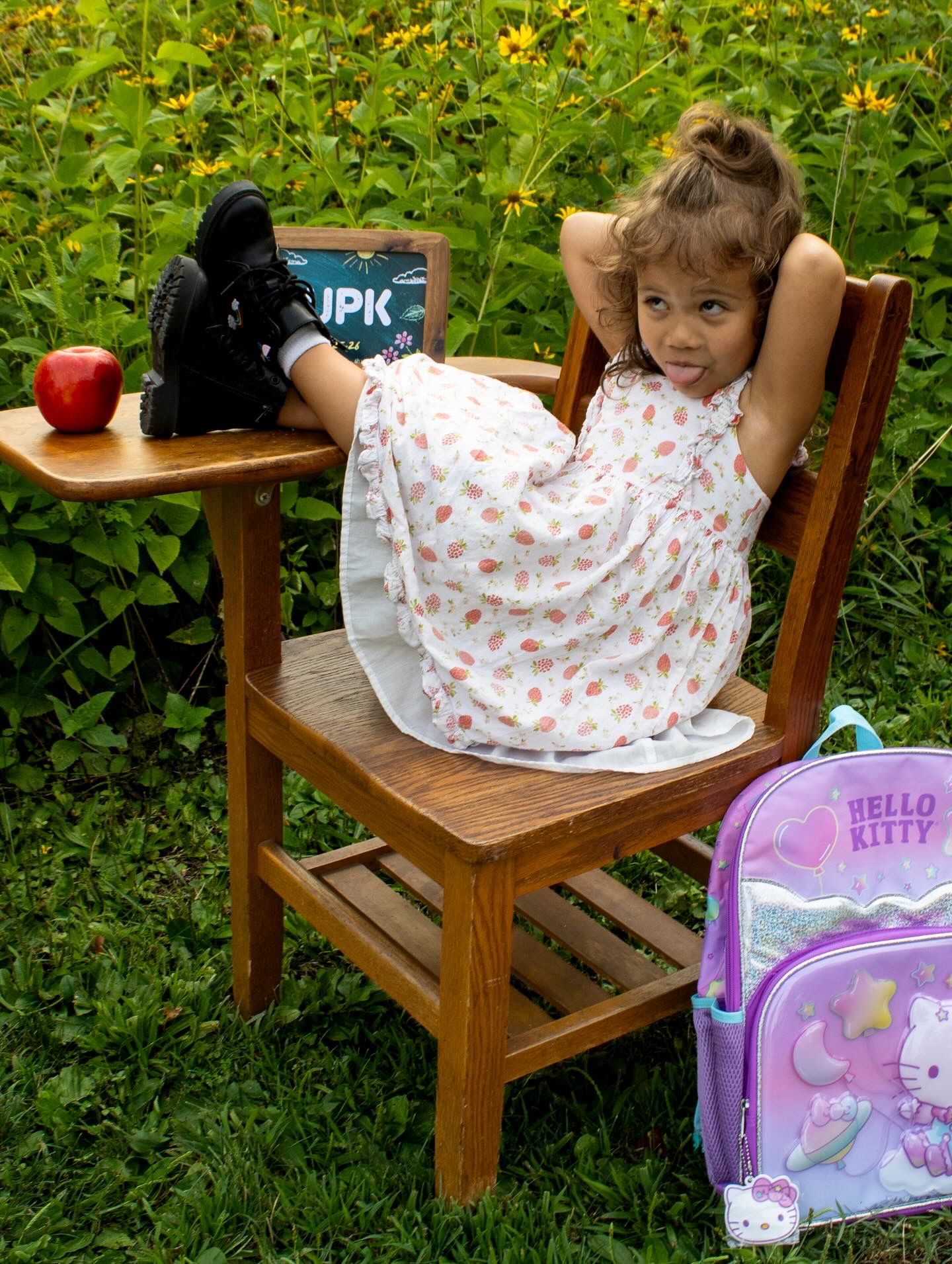 School photo setup with wooden chair, chalkboard sign and pink backpack in garden setting.