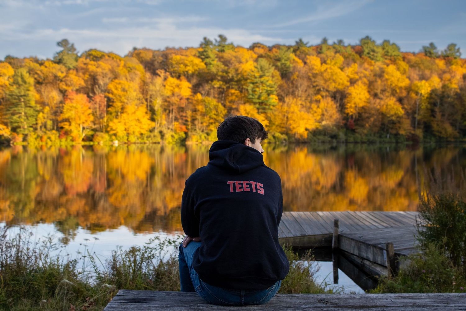 Person in black hoodie sits on dock overlooking lake with vibrant fall colors reflecting on water.