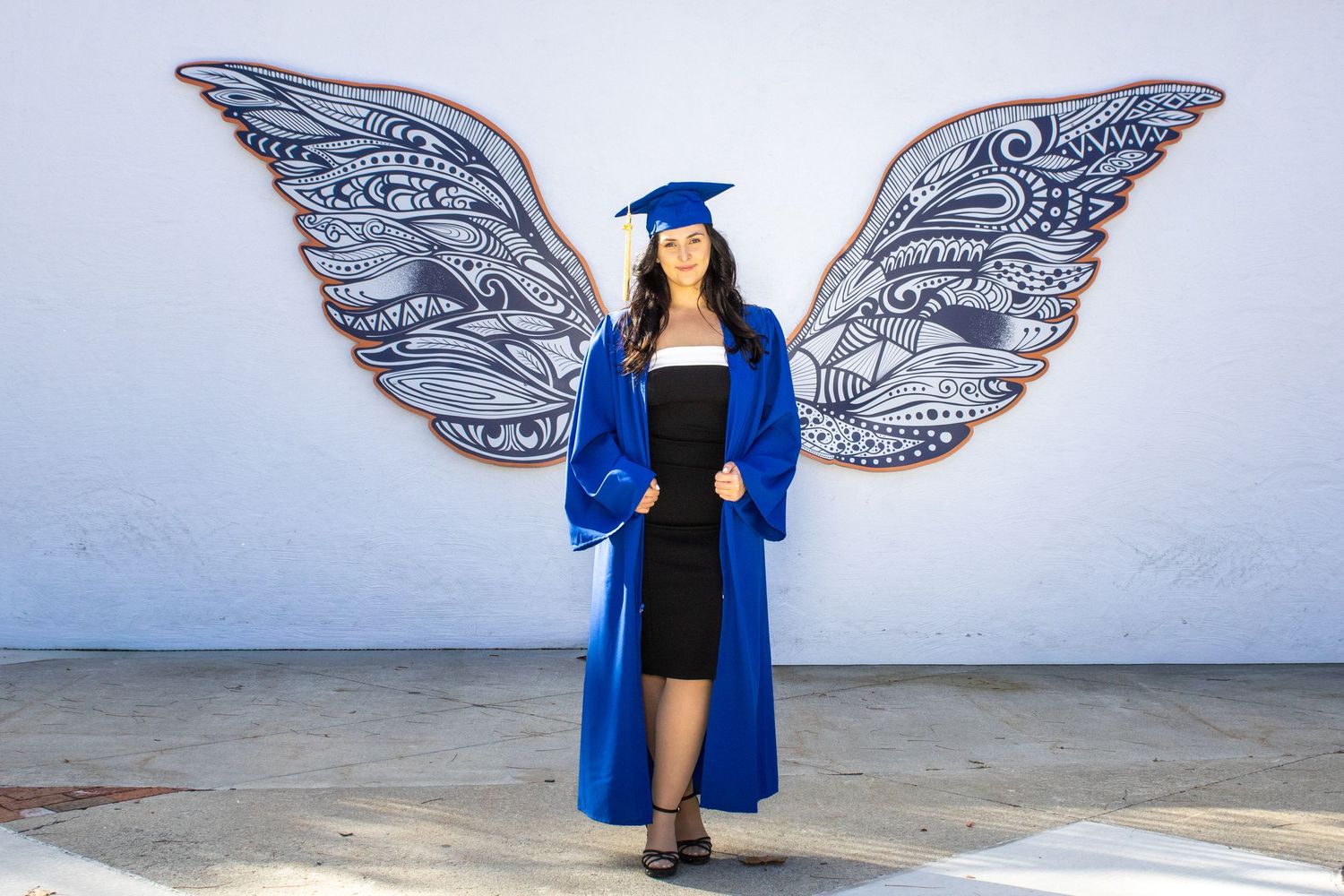 Graduate in blue regalia poses in front of decorative wing mural.