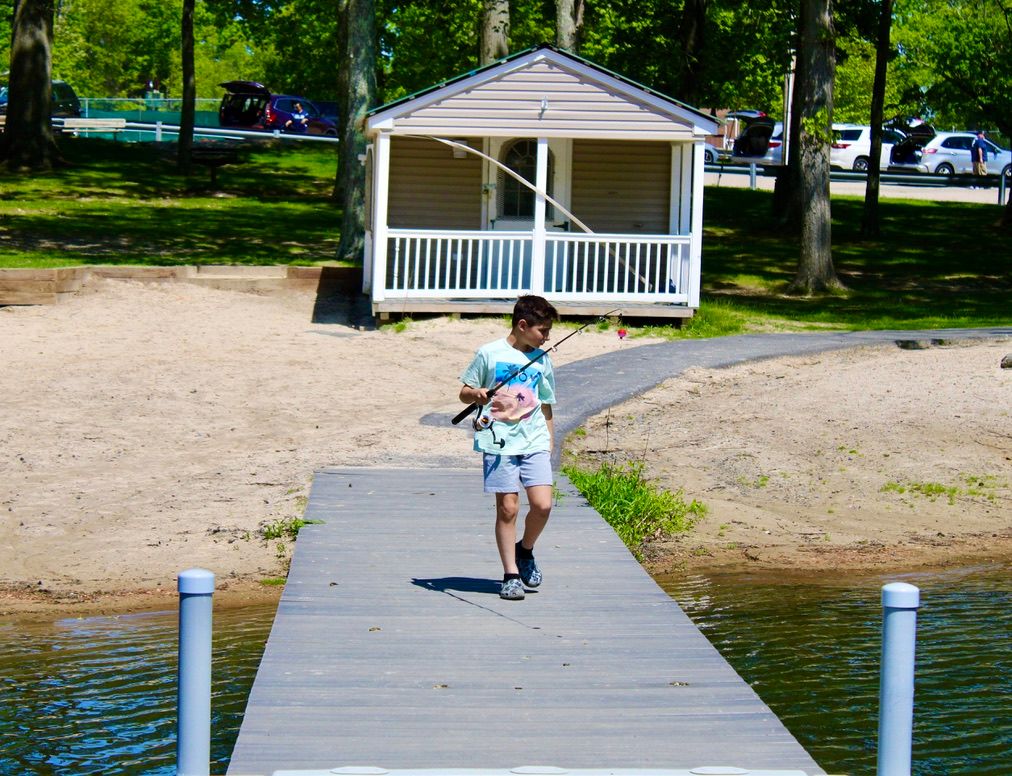A wooden dock extends into a calm lake surrounded by lush green trees and a white gazebo on the shore.