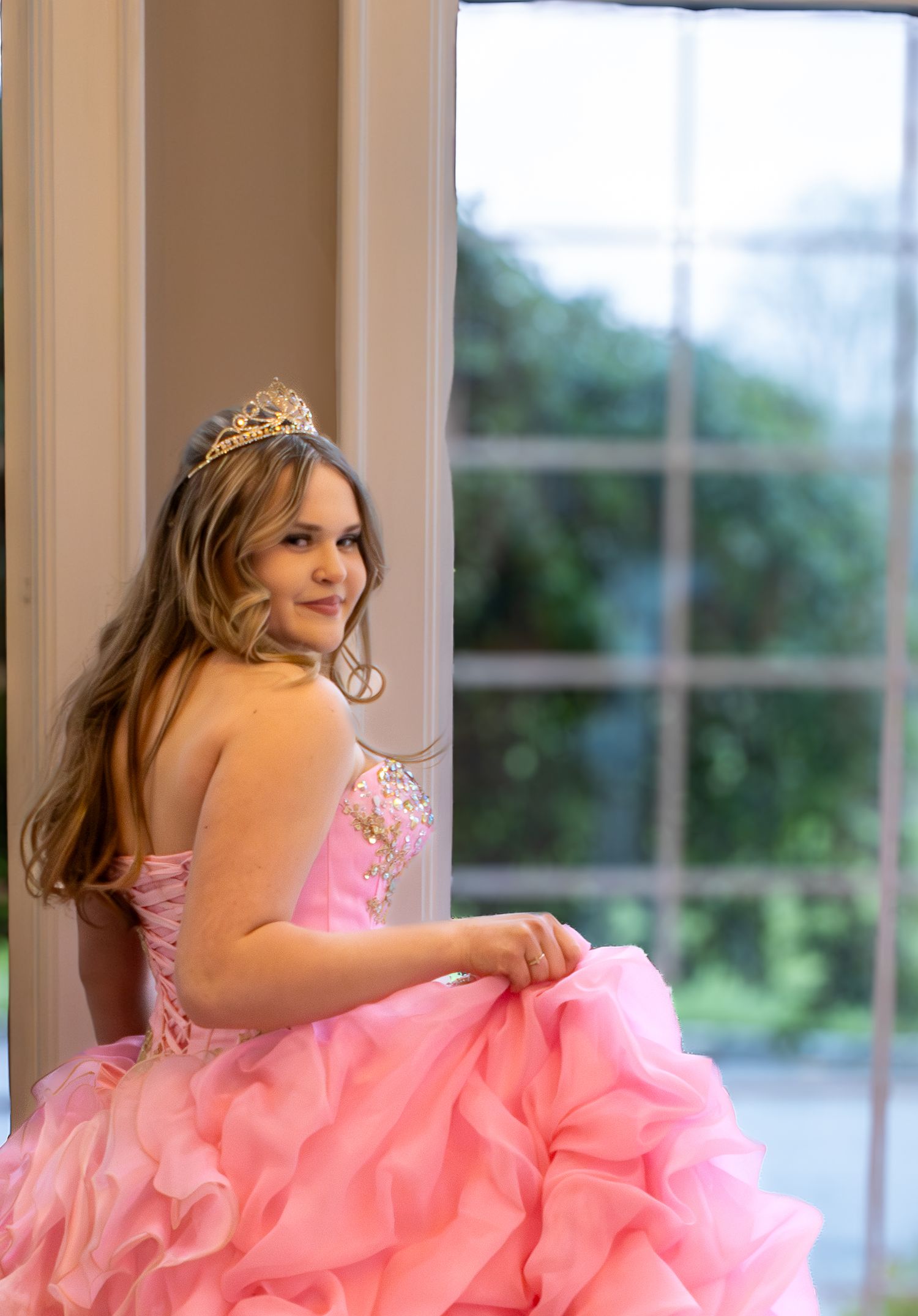 Young girl in pink ball gown and tiara poses gracefully by a large window for her quinceañera portrait.