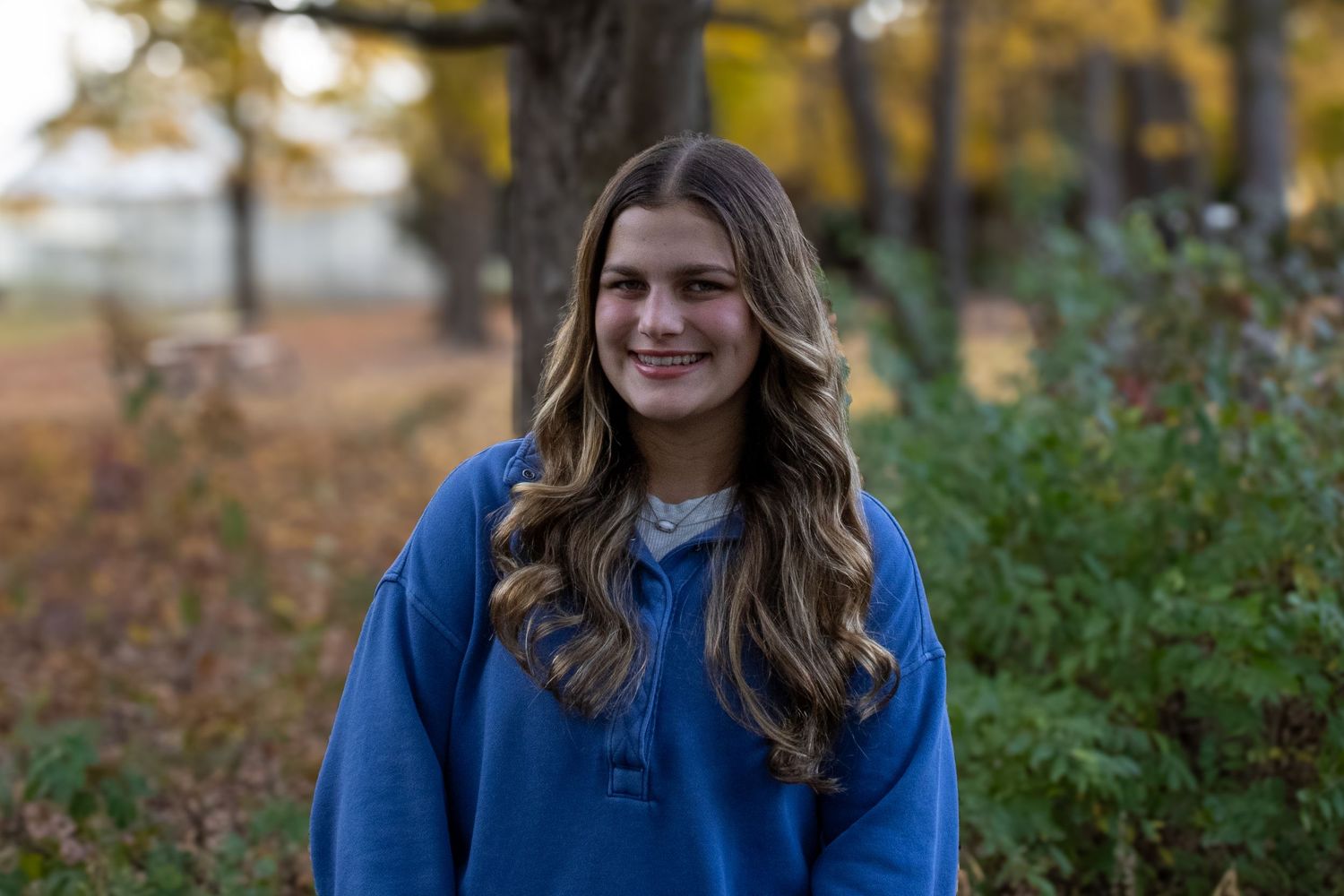 Young person in blue sweatshirt smiles outdoors during autumn season.