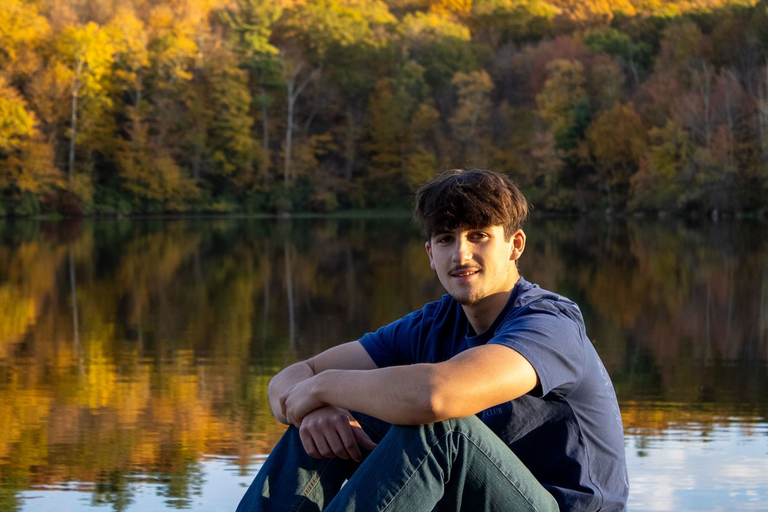 Person in casual clothing sits by serene lake with golden autumn foliage reflecting on water.