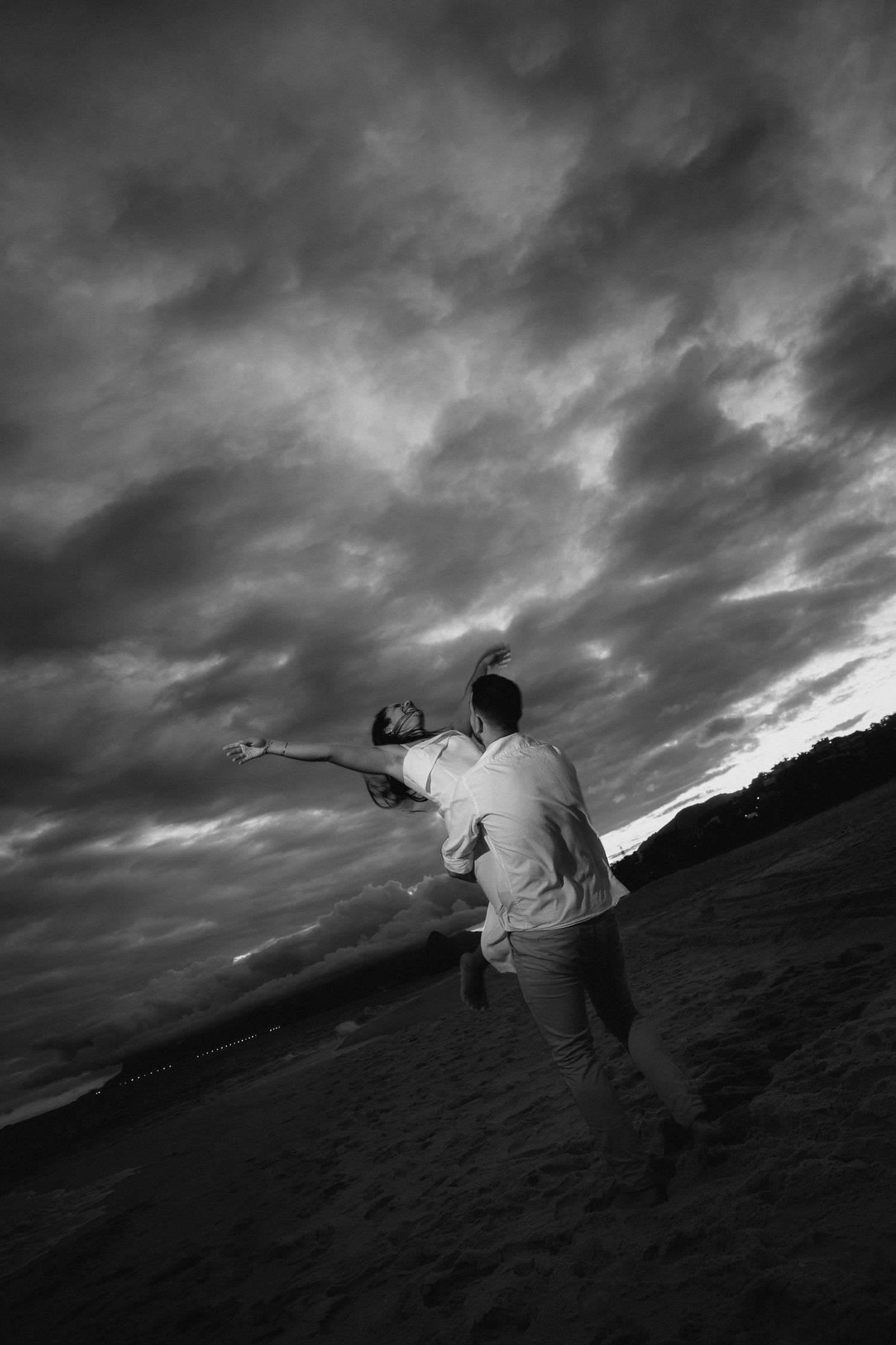 A silhueta de um casal está em uma encosta contra nuvens tempestuosas dramáticas em uma fotografia em preto e branco.
