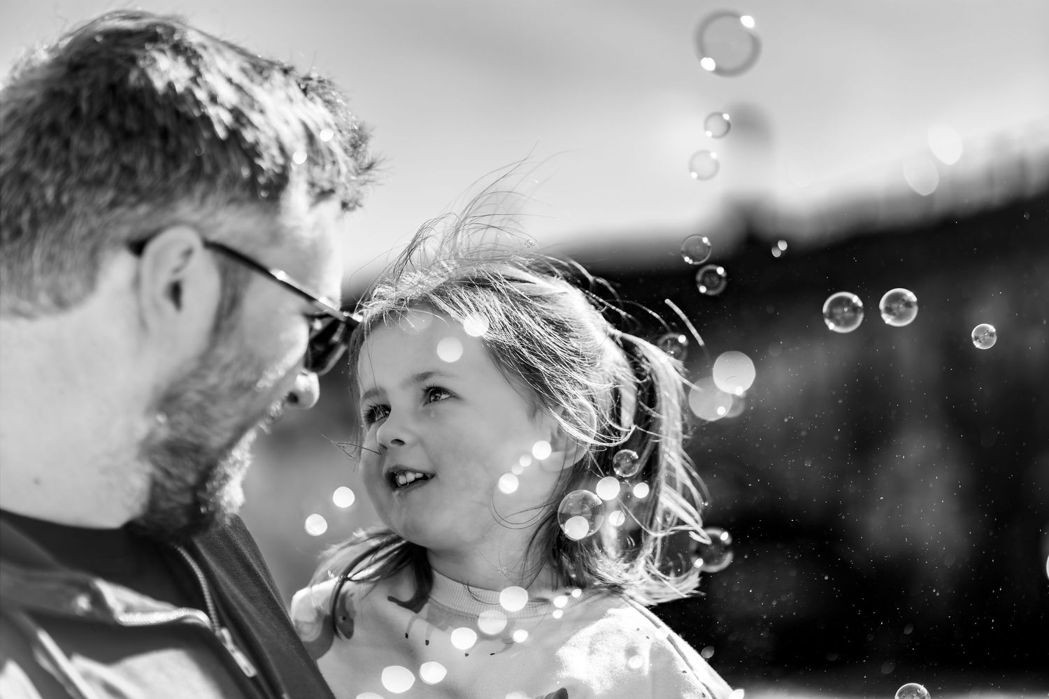A black and white image of a father and daughter looking into each other's eyes whilst bubbles are flying around them.