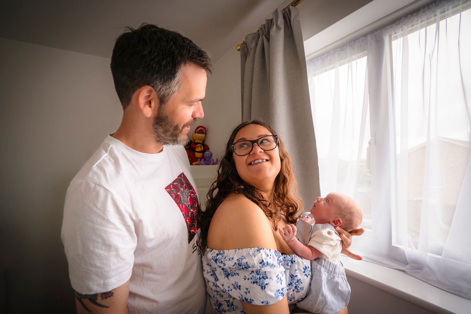 in home newborn session with a new mother and father, standing in front of a window and smiling at each other.