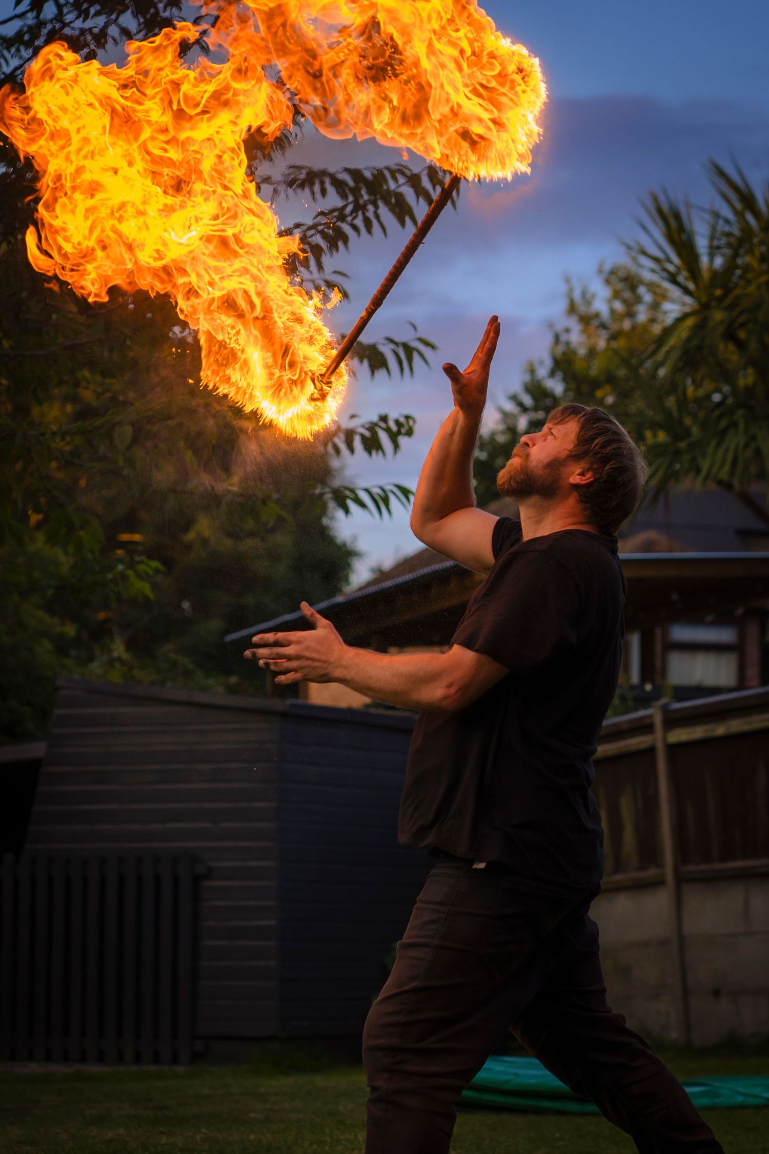 A bearded performer tossing a blazing fire staff skyward at dusk, flames curling bright against the trees and twilight.