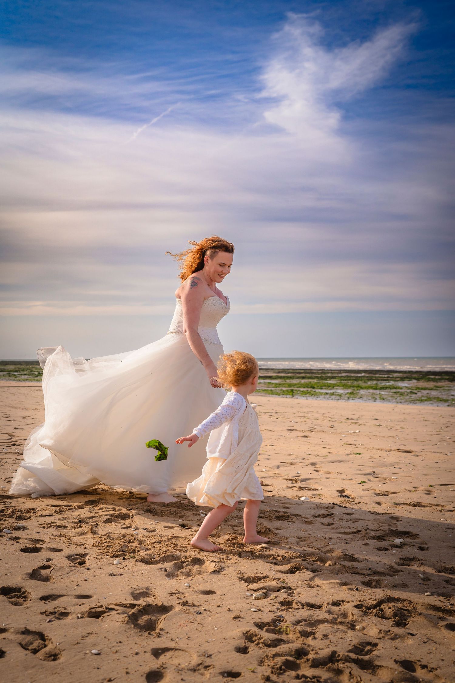 Mother and daughter on the beach walking towards the sea with a blue sky, white fluffy clouds and happy smiles.