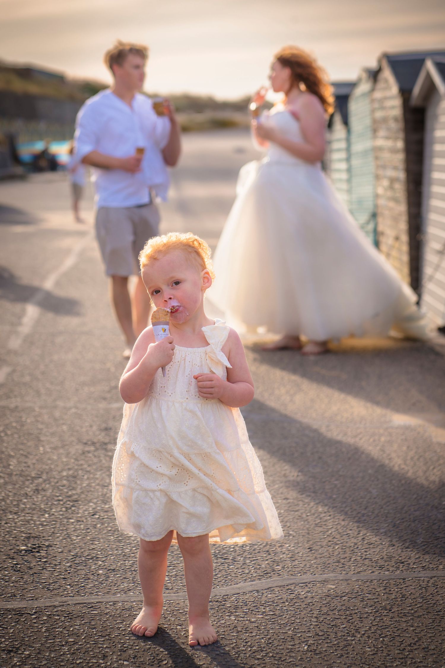 A little girl eating an ice-cream on a windy beach day with her parents out of focus behind her at a very special beach photo session.