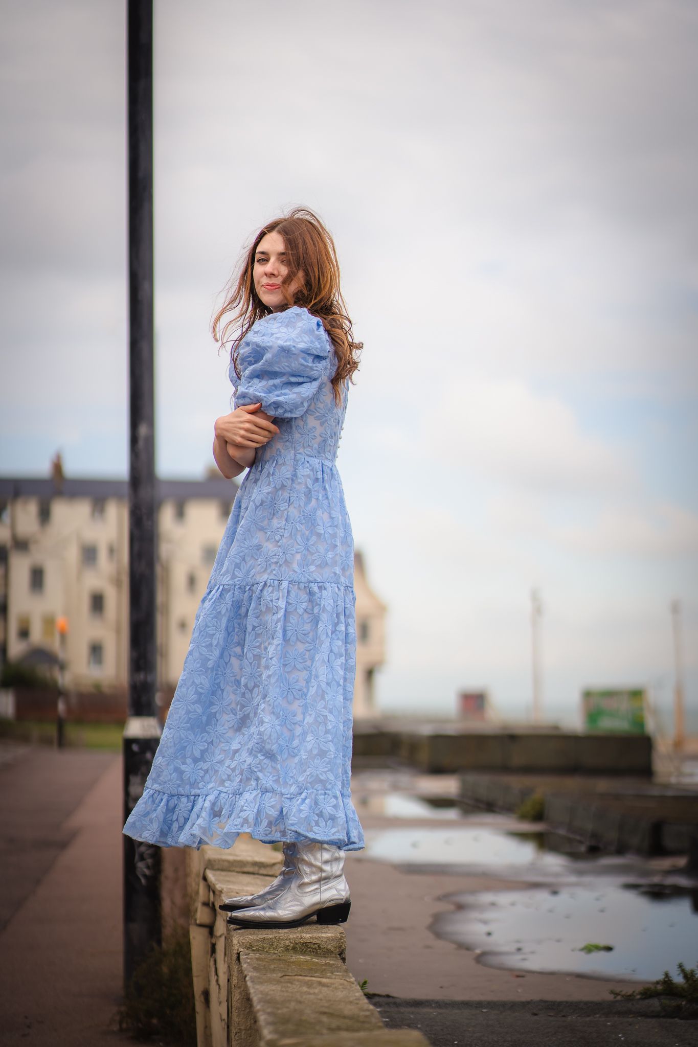 woman in a blue gown standing on an urban setting with the wind in her hair and her arms folded.