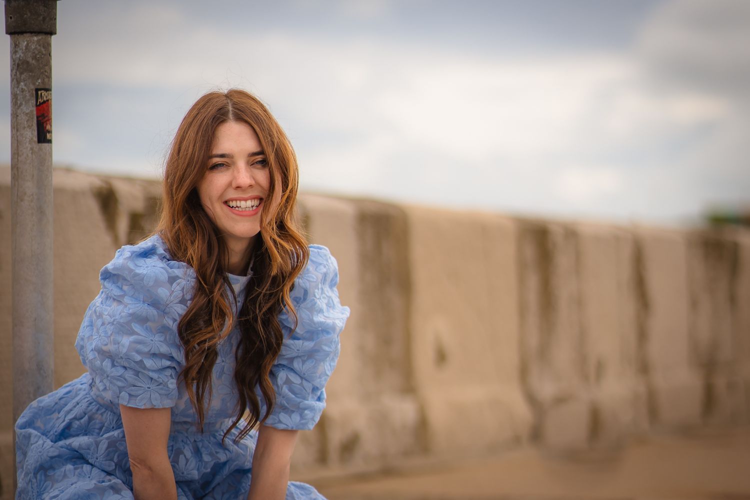 portrait of a woman with long hair and in a blue gown with the beach wall behind her, smiles and blue skies.
