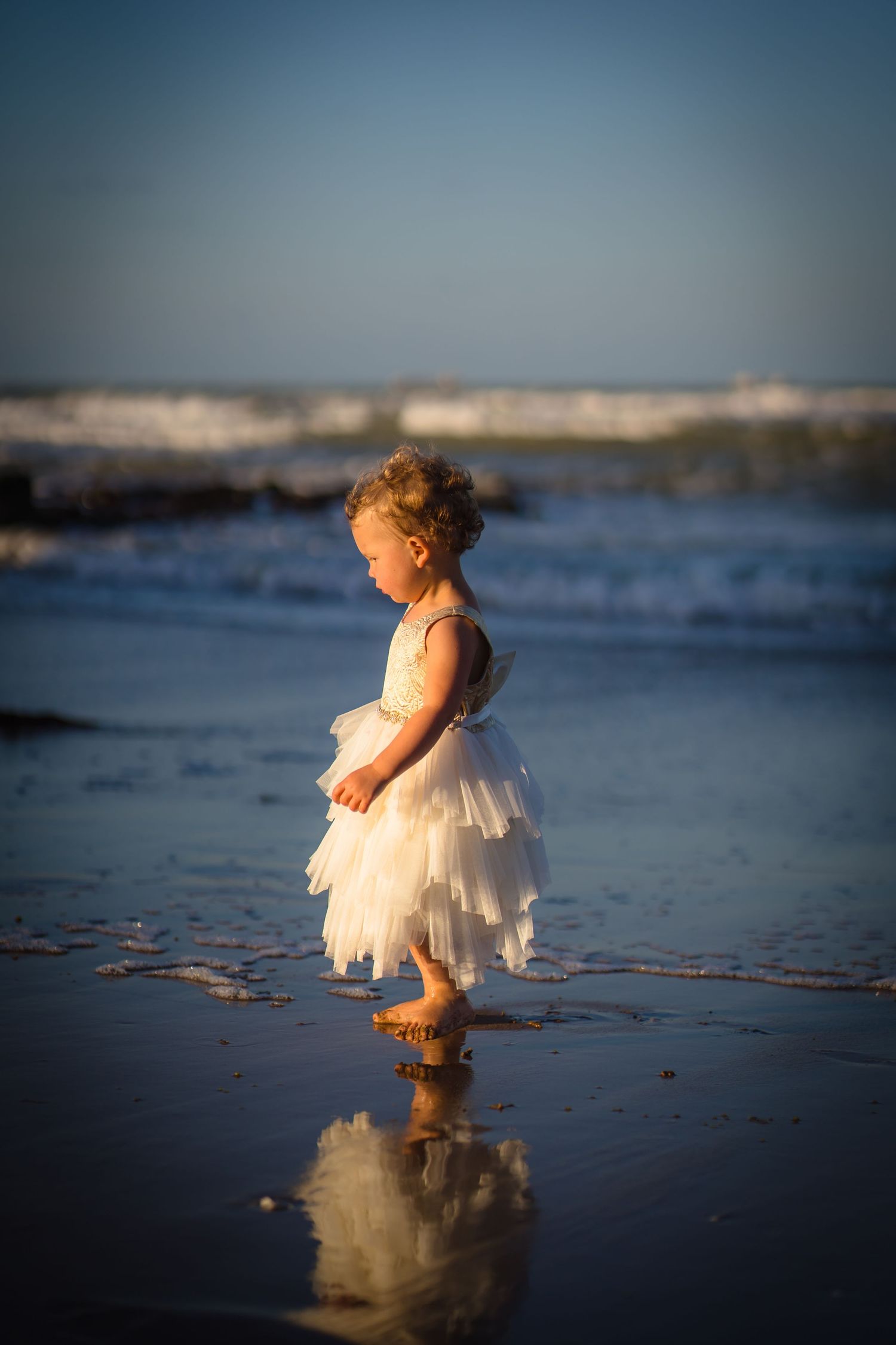 Beautiful toddler in a white gown standing on the beach with toes in the water and a reflection of the dress. Golden hour light and blue sea