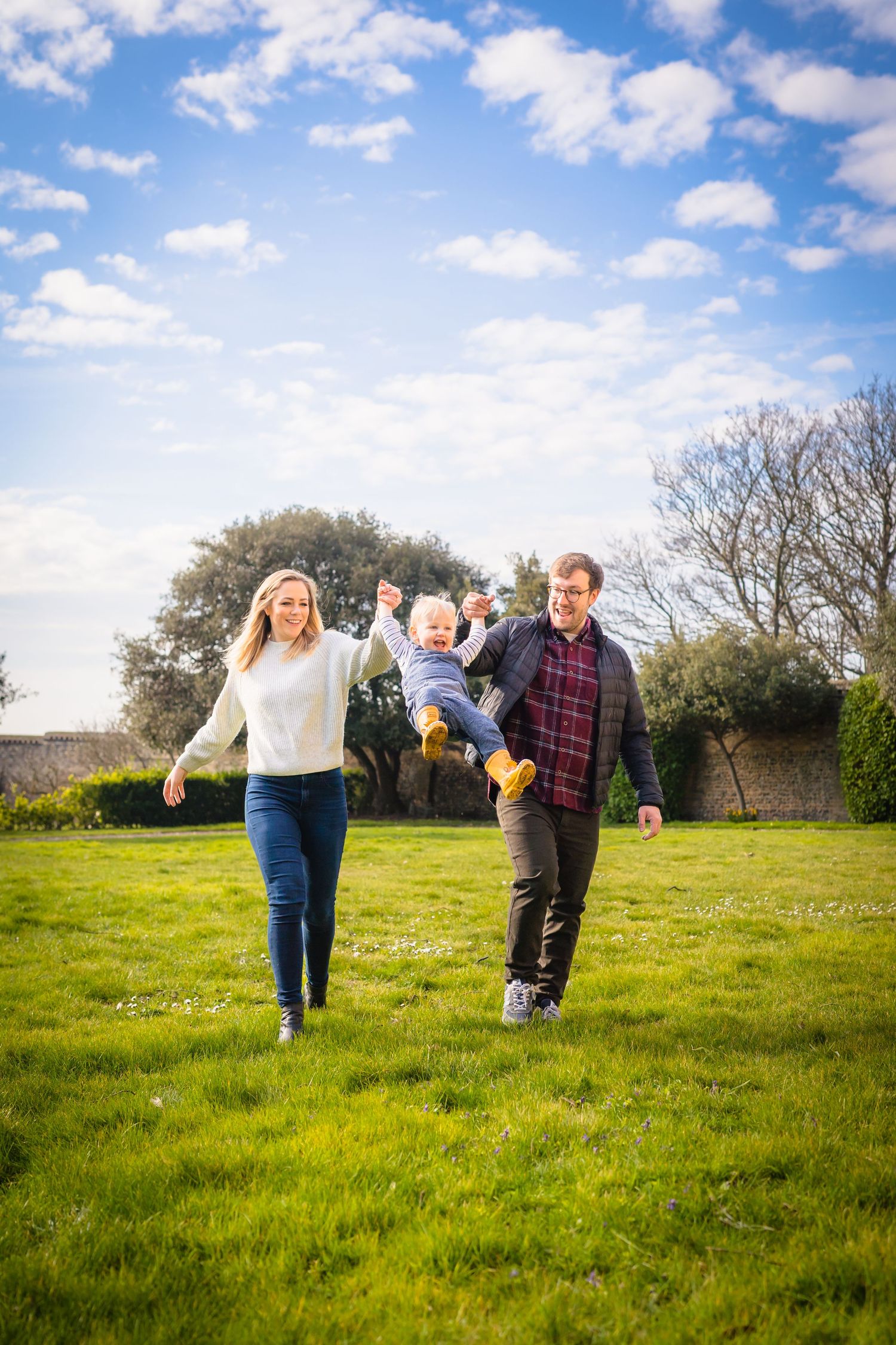 A mother and father swinging their son in the air whilst running through a Kent field on a beautiful summers day with blue sky and clouds.