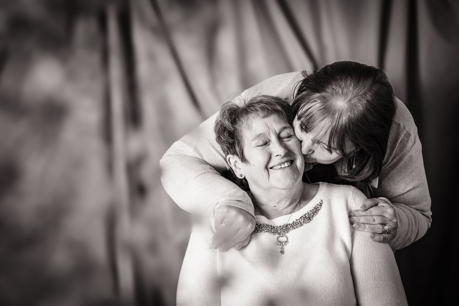 A mother and daughter sharing a kiss at an in home photo session, black and white, with out of focus flowers in front,  Shorely a memory.