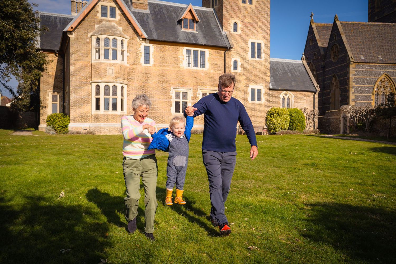 A grandmother and grandfather swinging their grandson in the air whilst running through a Kent field on a beautiful summers day