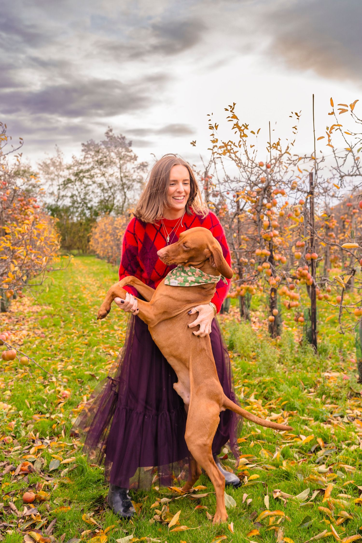 A lading in a red and purple outfit dancing with her orange dog in an autumnal apple orchard.
