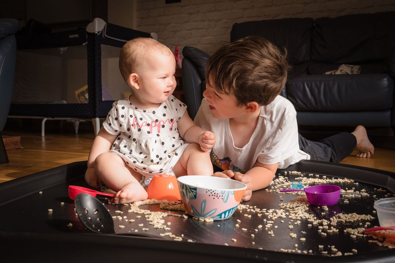 A brother playing with his baby sister in home, making a mess with rice and bowls.