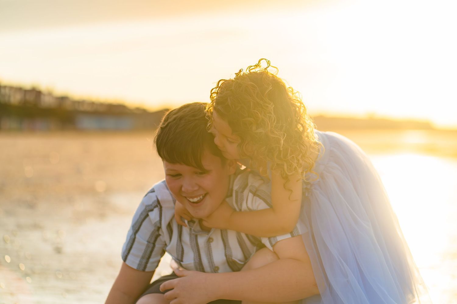 Hazy sunset photo of a sister hugging her big brother whilst on the beach with glowing sunlight around them.