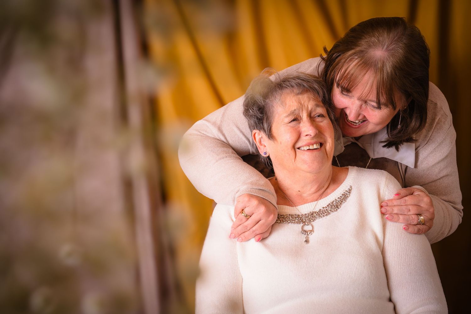 A mother and daughter sharing a joke at an in home photo session, browns and yellows with out of focus flowers in front,  Shorely a memory.