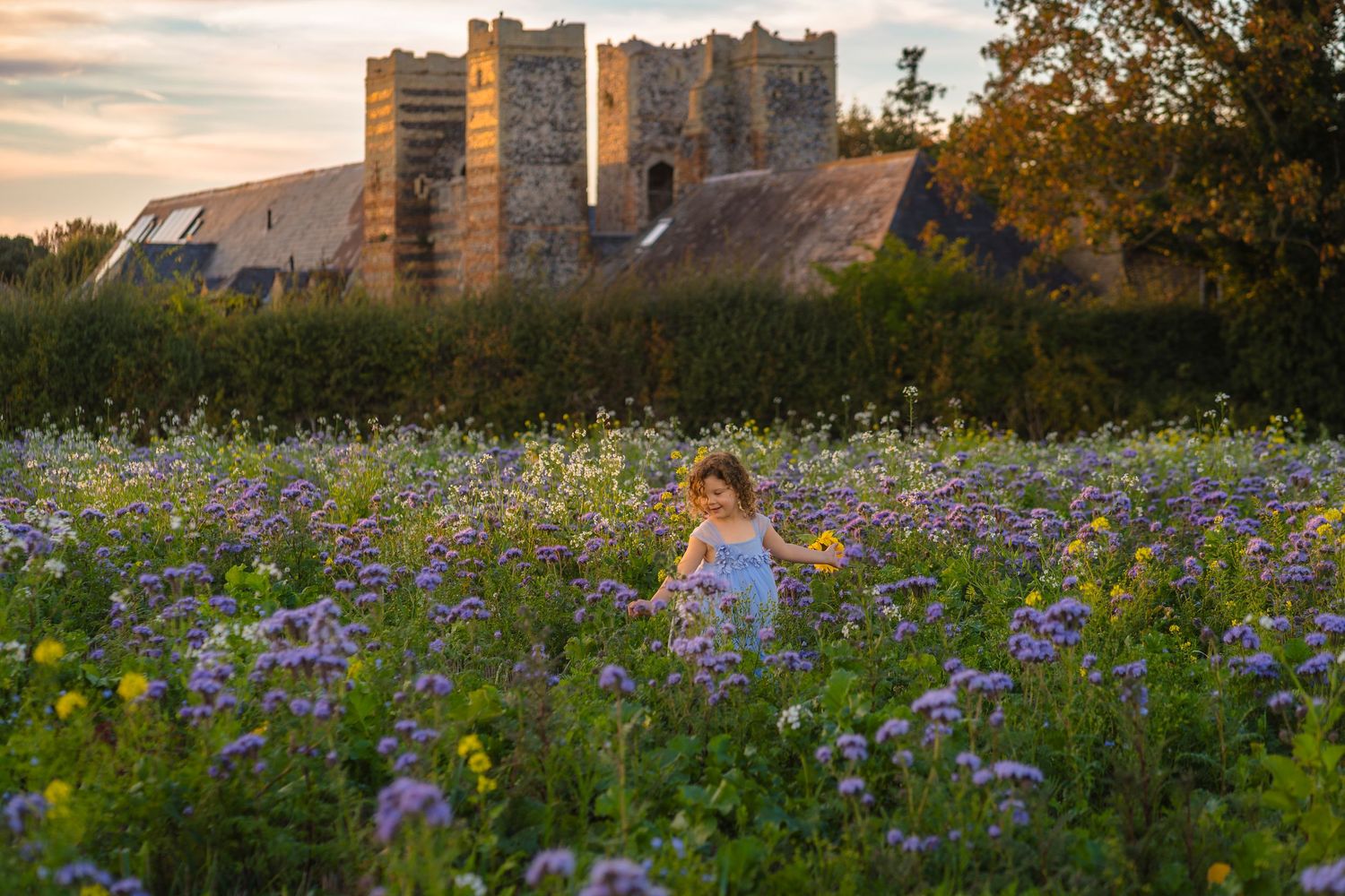 Beautiful portrait of a curly haired girl dancing through a flower field with a castle like building in the background Shorely a Memory Kent