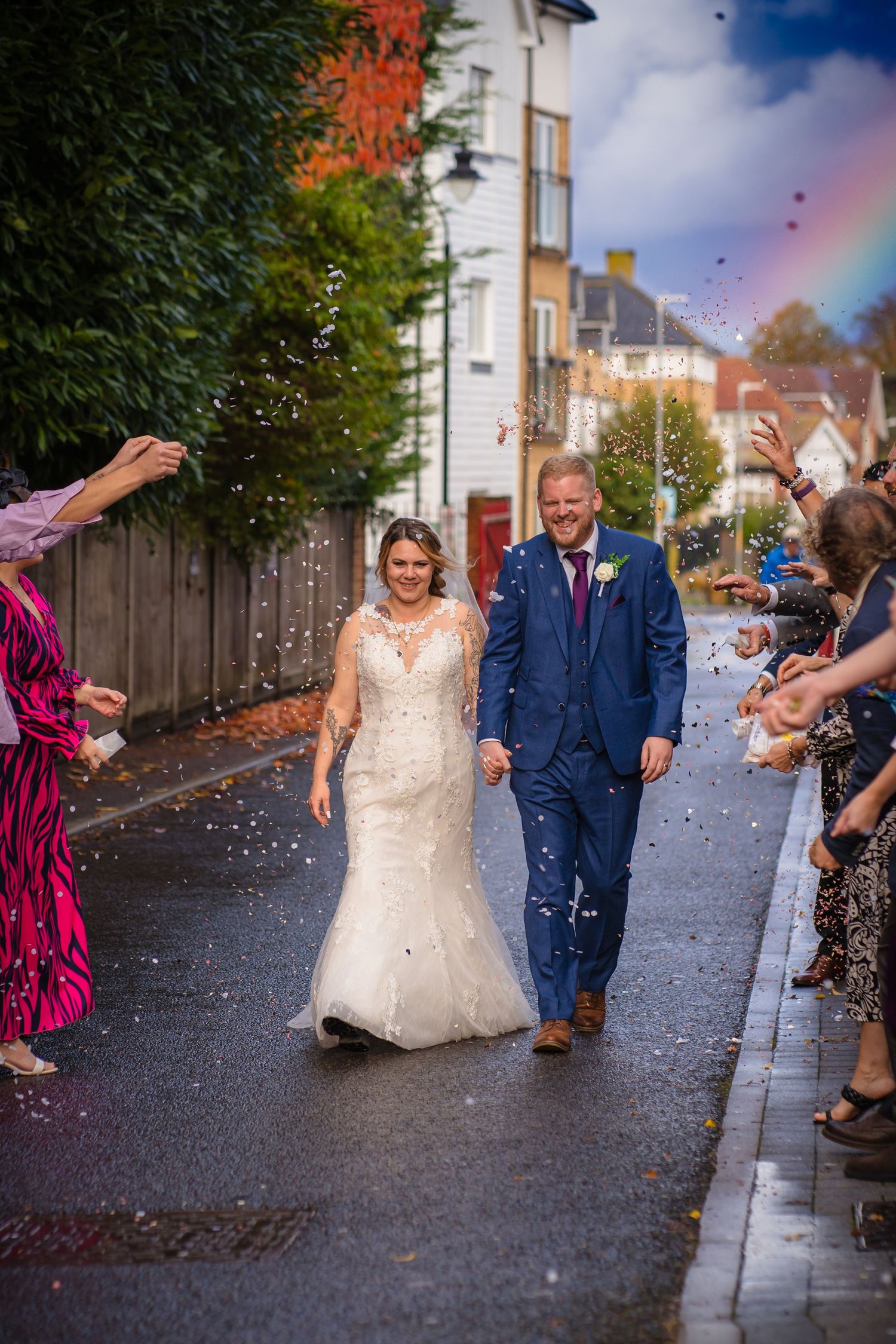 Bride and groom walking down a confetti tunnel with a rainbow in the sky behind them.