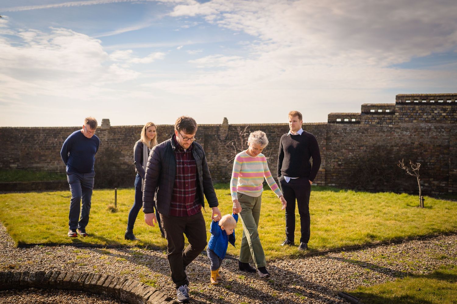 A candid family capture of an extended family walking across a garden together. Father and grandmother holding a toddler's hand.