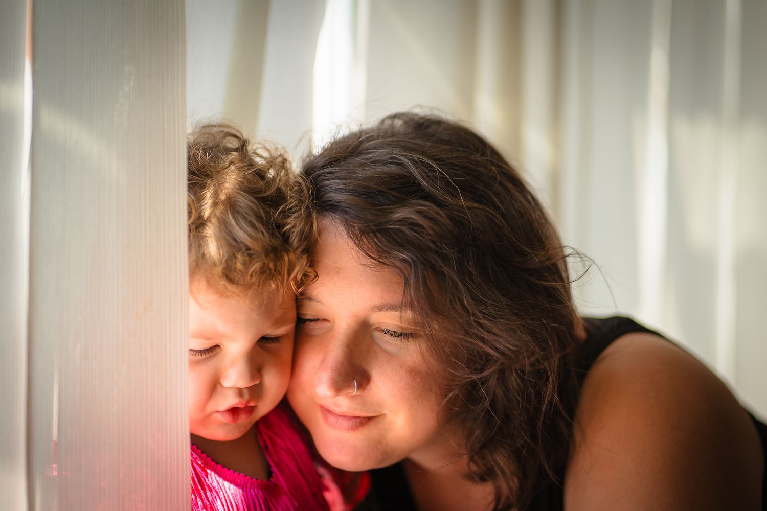 Mother and daughter in home near a window, both looking off camera as if reading together or sharing a moment,