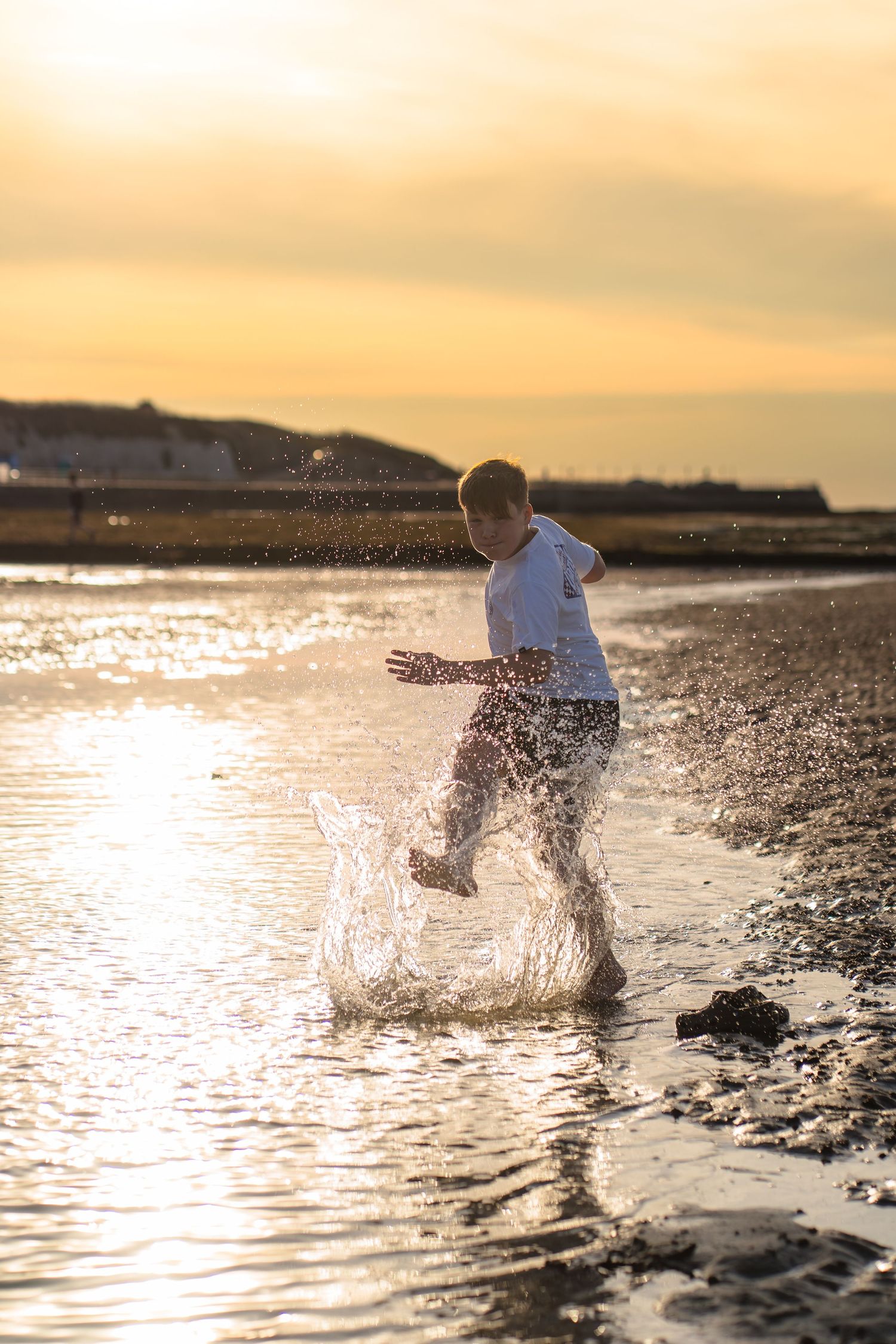 Sunset photo of a young boy kicking the water on the beach with orange skies behind and light highlighting the spaces.