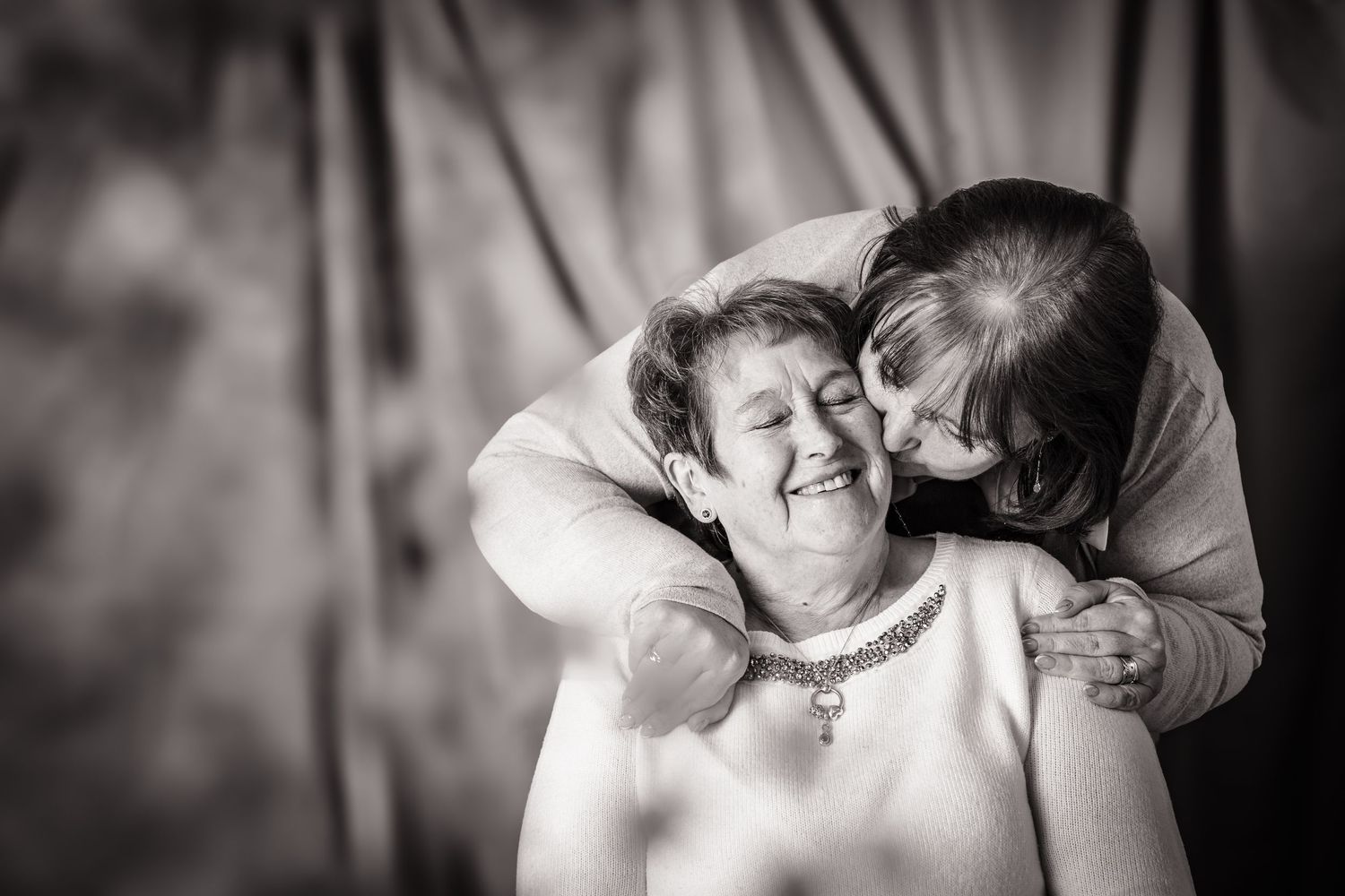 A mother and daughter sharing a kiss at an in home photo session, black and white, with out of focus flowers in front,  Shorely a memory.